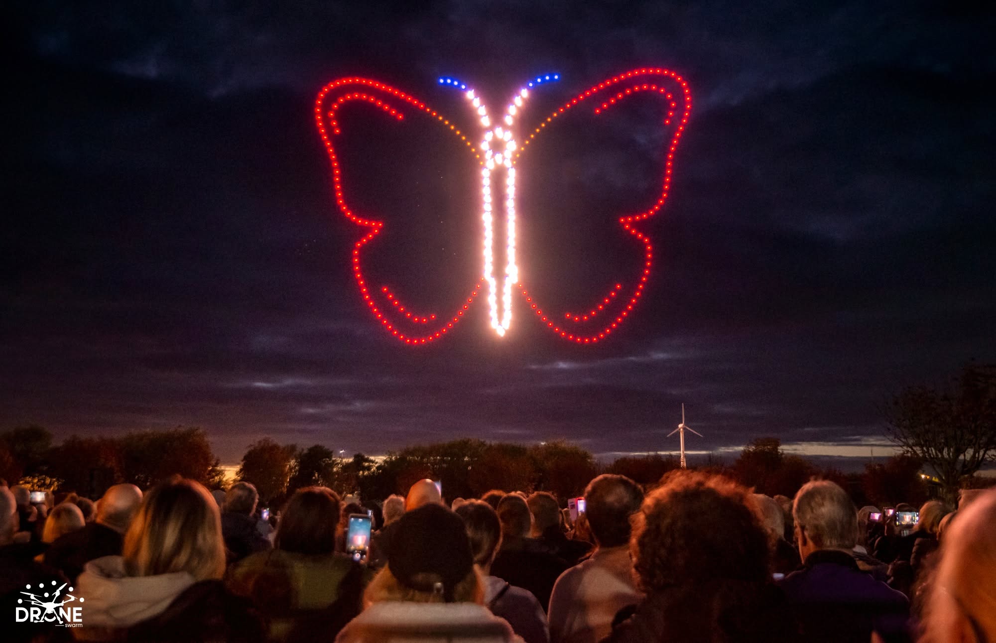 A brilliant red and blue butterfly drone formation with a sparkling centre above a watching crowd